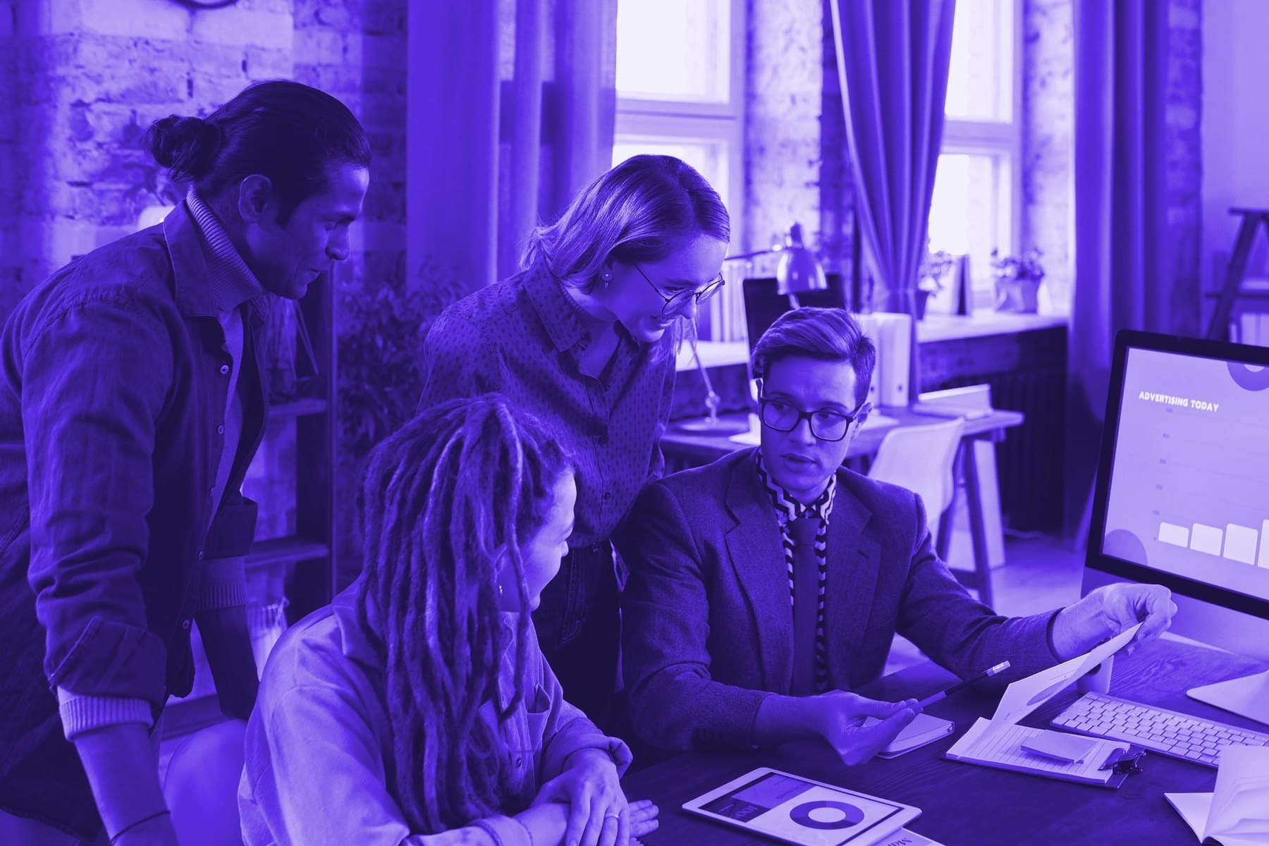 Four colleagues gathered at a desk, one pointing to a printed chart beside a monitor